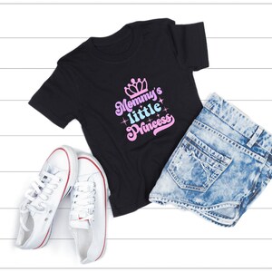 May include: Black t-shirt with the text "Mommy's little Princess" in pink and blue lettering with a crown above the text. The shirt is laid out on a white wooden surface with a pair of white sneakers and a pair of light blue denim shorts.