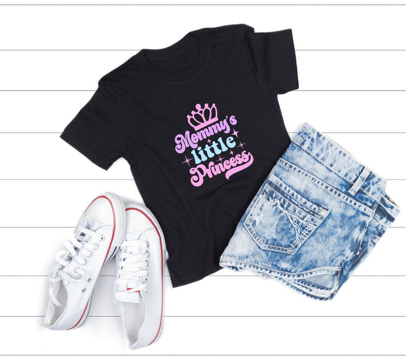 May include: Black t-shirt with the text "Mommy's little Princess" in pink and blue lettering with a crown above the text. The shirt is laid out on a white wooden surface with a pair of white sneakers and a pair of light blue denim shorts.