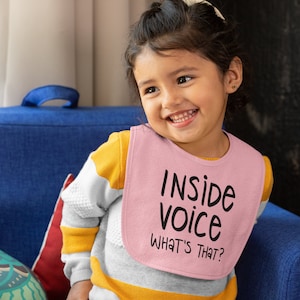 May include: A young child wearing a pink bib with the text "Inside voice What's that?" printed on it. The child is sitting on a blue sofa and smiling.
