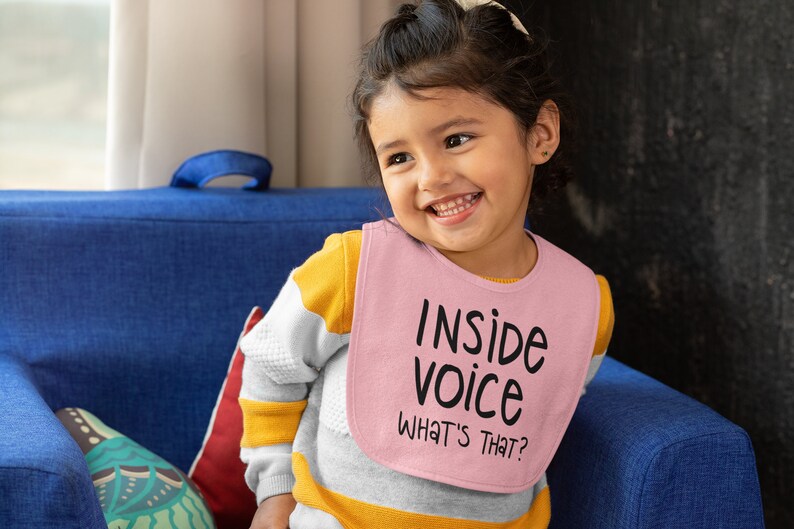 May include: A young child wearing a pink bib with the text "Inside voice What's that?" printed on it. The child is sitting on a blue sofa and smiling.