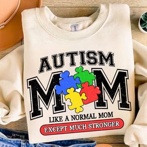 May include: A cream colored sweatshirt with the text "AUTISM MOM" in black letters with a vintage style. The "M" is made up of colorful puzzle pieces. The text "LIKE A NORMAL MOM EXCEPT MUCH STRONGER" is printed in red below the main text.