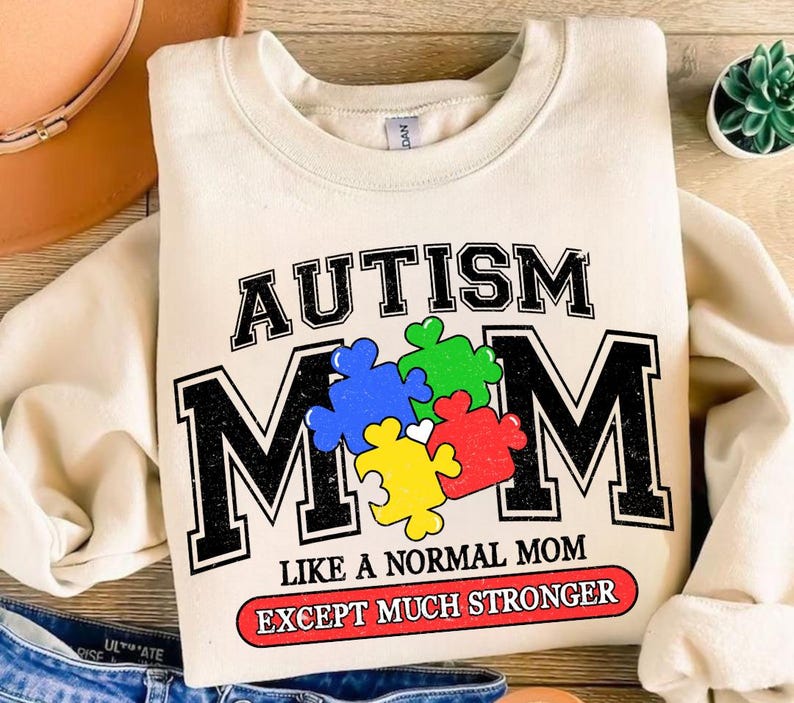 May include: A cream colored sweatshirt with the text "AUTISM MOM" in black letters with a vintage style. The "M" is made up of colorful puzzle pieces. The text "LIKE A NORMAL MOM EXCEPT MUCH STRONGER" is printed in red below the main text.
