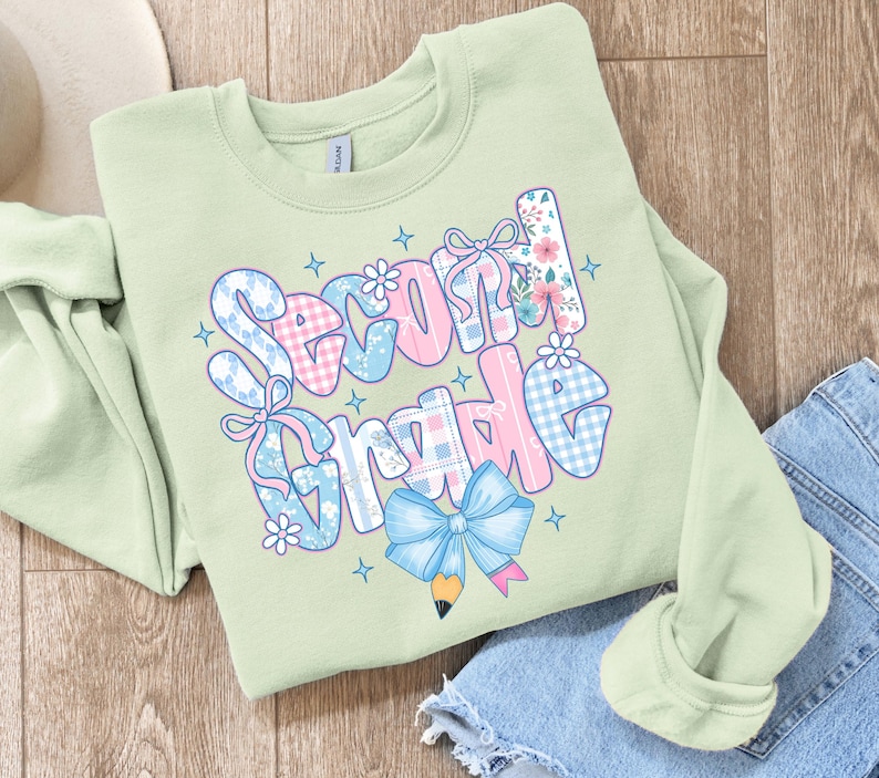 Może przedstawiać: Jasnozielona bluza z napisem "Second Grade" w kolorowym, fantazyjnym wzorze. Tekst zawiera r&oacute;żowe, niebieskie i białe wzory, kokardy i kwiatowe akcenty, z niebieską grafiką oł&oacute;wka i kokardy.