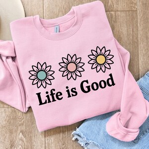 May include: A pale pink sweatshirt featuring three daisy designs and the text "Life is Good" in black. The daisies have teal, pink, and yellow centres. A pair of blue denim shorts and a white hat are also in the image.