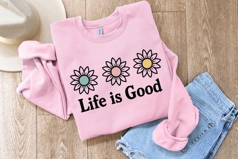 May include: A pale pink sweatshirt featuring three daisy designs and the text "Life is Good" in black. The daisies have teal, pink, and yellow centres. A pair of blue denim shorts and a white hat are also in the image.