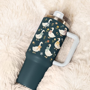May include: A teal tumbler with a white handle and straw. The tumbler is decorated with a pattern of white geese wearing colorful bows and surrounded by small flowers. The lid is silver.