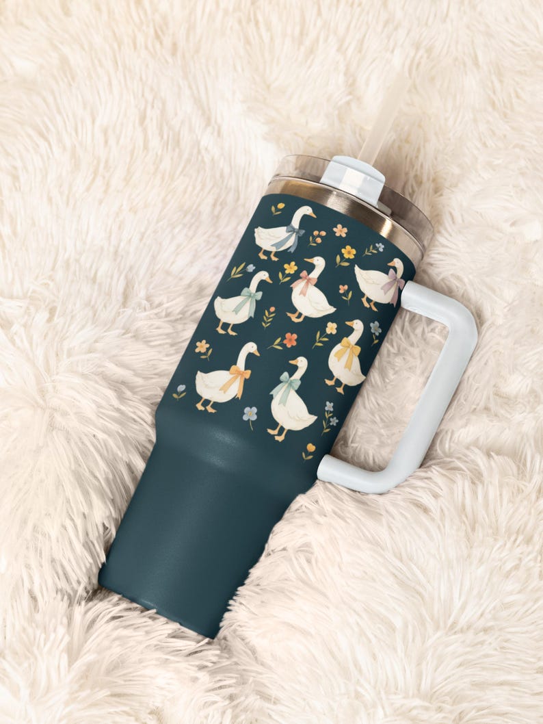 May include: A teal tumbler with a white handle and straw. The tumbler is decorated with a pattern of white geese wearing colorful bows and surrounded by small flowers. The lid is silver.