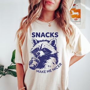Puede incluir: Camiseta color crema con un gráfico de mapache azul marino sosteniendo una bebida. La palabra "SNACKS" está encima del mapache, y "MAKE ME NICER" está debajo. La camiseta tiene mangas cortas y un corte holgado.