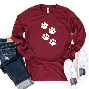 May include: A maroon long-sleeved shirt with five white paw prints on the front. The shirt is paired with blue jeans and white sneakers.