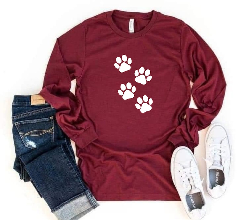 May include: A maroon long-sleeved shirt with five white paw prints on the front. The shirt is paired with blue jeans and white sneakers.
