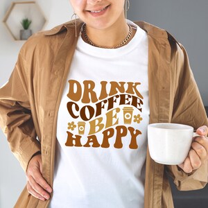 May include: A white t-shirt with the text "Drink Coffee Be Happy" in brown and orange letters with a retro font. The text is surrounded by coffee cup illustrations.
