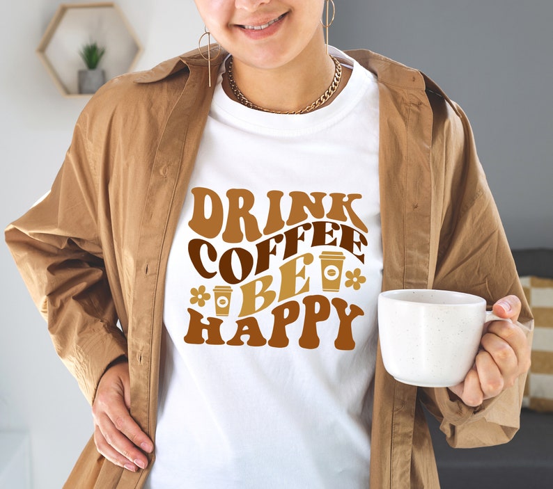May include: A white t-shirt with the text "Drink Coffee Be Happy" in brown and orange letters with a retro font. The text is surrounded by coffee cup illustrations.