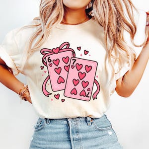 May include: Cream-colored t-shirt featuring a graphic of two pink playing cards with heart symbols and the numbers 6 and 7. The cards are tied with a pink ribbon and surrounded by small hearts. A cute and romantic design.