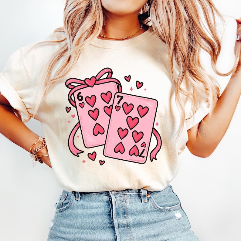 May include: Cream-colored t-shirt featuring a graphic of two pink playing cards with heart symbols and the numbers 6 and 7. The cards are tied with a pink ribbon and surrounded by small hearts. A cute and romantic design.