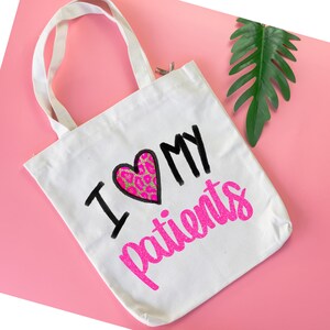 Puede incluir: Bolsa de tela blanca con un dise&ntilde;o de purpurina rosa que dice "Amo a mis pacientes" con un coraz&oacute;n rosa y dorado.
