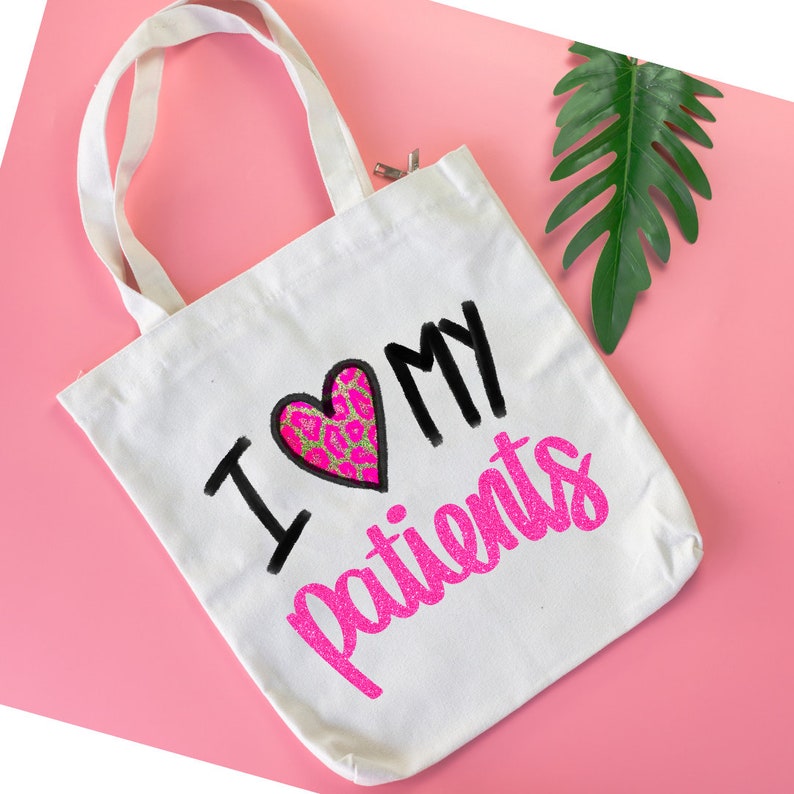 Puede incluir: Bolsa de tela blanca con un dise&ntilde;o de purpurina rosa que dice "Amo a mis pacientes" con un coraz&oacute;n rosa y dorado.