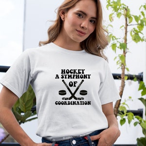 May include: White t-shirt with black text that reads "Hockey A Symphony Of Coordination" with a graphic of two hockey sticks crossed over a puck.