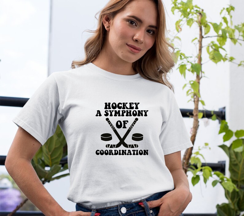 May include: White t-shirt with black text that reads "Hockey A Symphony Of Coordination" with a graphic of two hockey sticks crossed over a puck.