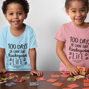 May include: Two children wearing light blue and pink t-shirts with the text "100 Days of Livin' That Kindergarten Life" printed on them. They are playing a game with cards featuring cartoon characters.