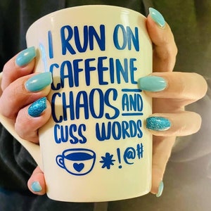 May include: White ceramic mug with blue lettering that reads "I run on caffeine chaos and cuss words *!@#" with a small blue coffee cup with a heart on it. The mug is being held by a hand with blue nail polish.