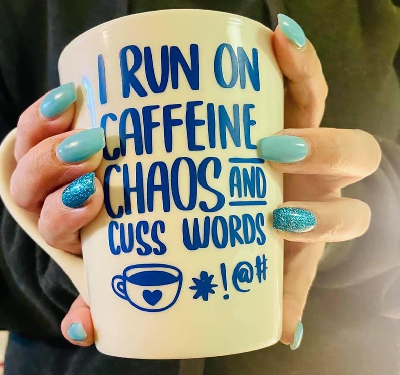 May include: White ceramic mug with blue lettering that reads "I run on caffeine chaos and cuss words *!@#" with a small blue coffee cup with a heart on it. The mug is being held by a hand with blue nail polish.