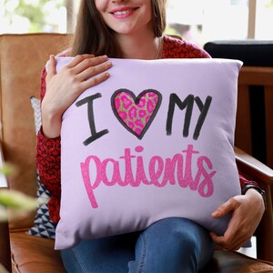 Puede incluir: Una almohada de color morado con el texto "Amo a mis pacientes" en letras negras y rosas con purpurina. Un coraz&oacute;n rosa con un dise&ntilde;o de estampado de leopardo est&aacute; en el centro de la almohada.