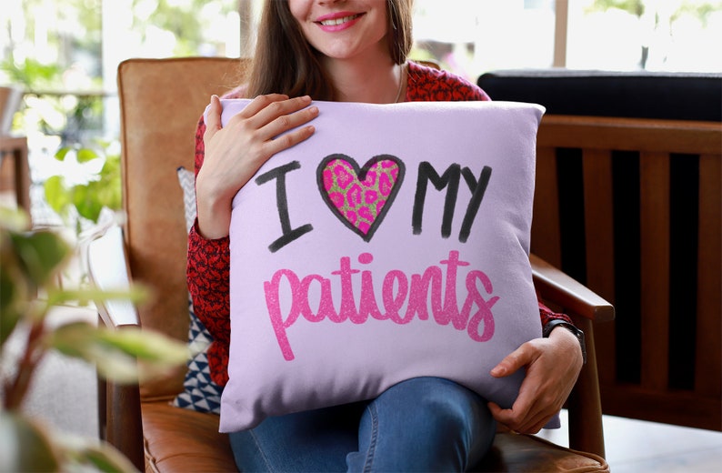 Puede incluir: Una almohada de color morado con el texto "Amo a mis pacientes" en letras negras y rosas con purpurina. Un coraz&oacute;n rosa con un dise&ntilde;o de estampado de leopardo est&aacute; en el centro de la almohada.