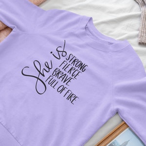 May include: A lavender sweatshirt with the text "She is strong, fierce, brave, full of fire" printed in black.