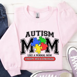 May include: Pink sweatshirt with a graphic that says "Autism Mom Like a Normal Mom Except Much Stronger". The graphic features a puzzle piece design in blue, green, yellow, and red.