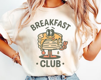 Könnte beinhalten: Cremefarbenes Sweatshirt mit der Aufschrift "BREAKFAST CLUB" über und unter einem Cartoon-Pfannkuchenstapel mit Sirup, Augen und einem Friedenszeichen. Die Pfannkuchen haben Beine und Arme. Das Shirt wird von einer Person getragen.