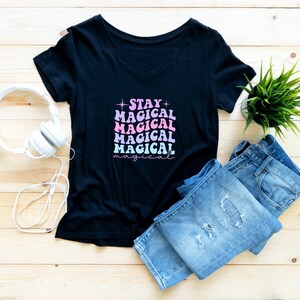 May include: Black v-neck t-shirt with the text "STAY MAGICAL" in pink and blue lettering. The shirt is laid flat on a wooden surface with a pair of blue jeans and white headphones.