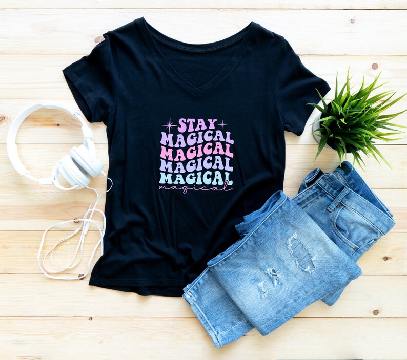 May include: Black v-neck t-shirt with the text "STAY MAGICAL" in pink and blue lettering. The shirt is laid flat on a wooden surface with a pair of blue jeans and white headphones.