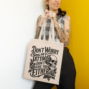 May include: Beige tote bag with the text "Don't Worry My Tattoos Don't Like You Either" in black. The design includes a skull and roses. The bag is held by a person wearing a black and white checkered top and black pants.