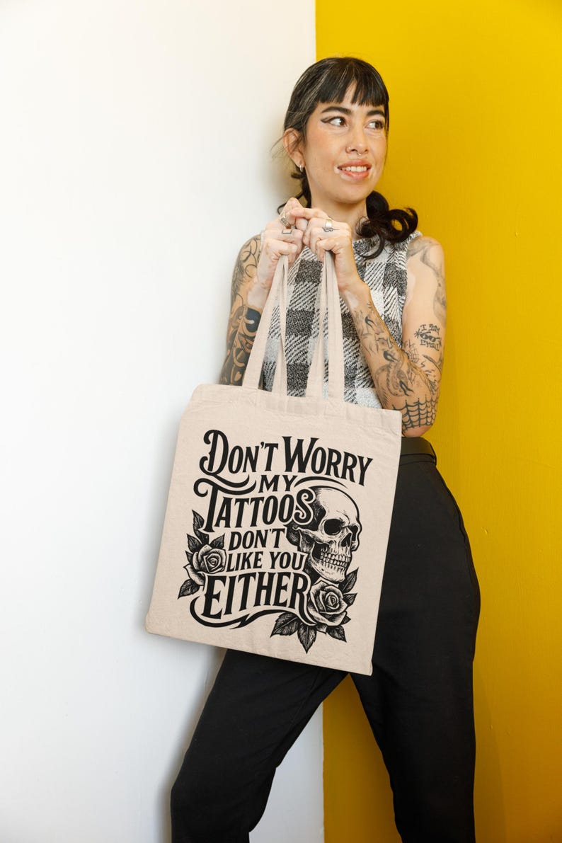 May include: Beige tote bag with the text "Don't Worry My Tattoos Don't Like You Either" in black. The design includes a skull and roses. The bag is held by a person wearing a black and white checkered top and black pants.