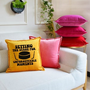 May include: A yellow throw pillow with the text "Setting the stage for unforgettable moments" and a black hockey puck graphic. The pillow is on a white couch with pink and red throw pillows.