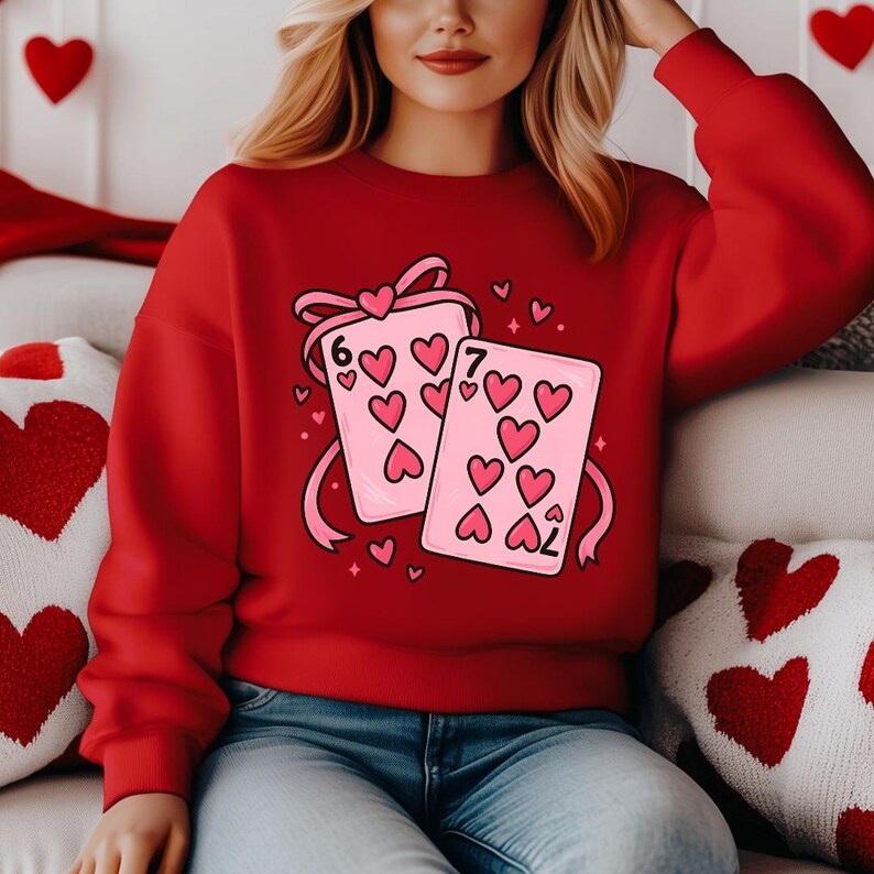 May include: A red sweatshirt featuring a graphic of two playing cards with heart suit symbols. The cards are pink with the numbers 6 and 7. Pink hearts and a pink ribbon complete the design. The sweatshirt is a Valentine's Day theme.