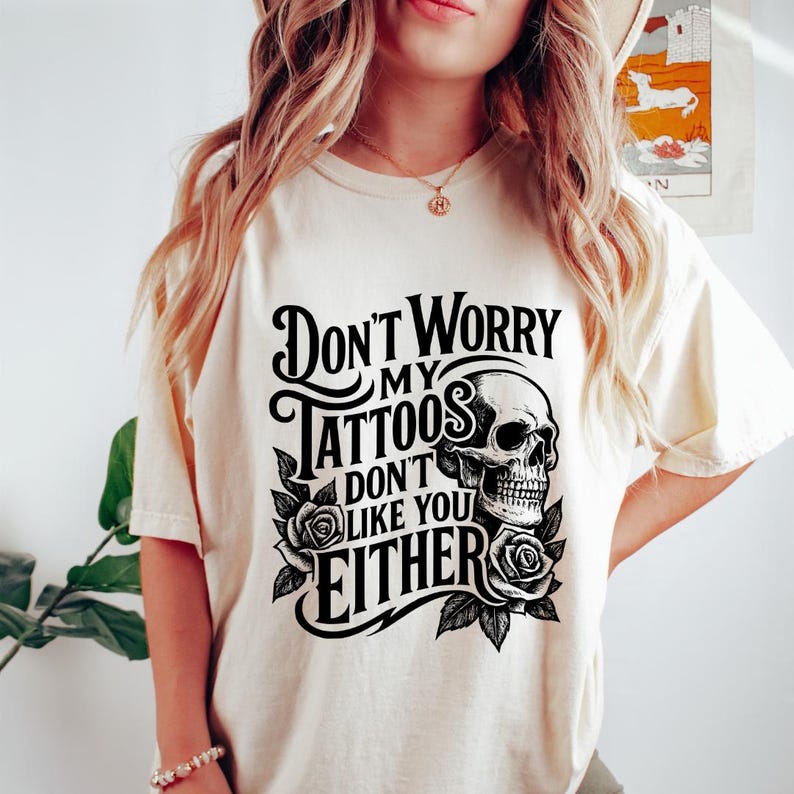 May include: Cream-colored t-shirt featuring the text "Don't Worry My Tattoos Don't Like You Either" in a black, gothic-style font. A skull and roses graphic is centered below the text.