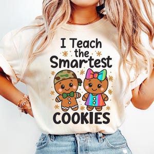 May include: A cream-coloured t-shirt with the words "I Teach the Smartest COOKIES." The design shows two gingerbread cookie characters, one with a camouflage hat and bow tie, the other with a rainbow bow and dress. Snowflake accents are also included.