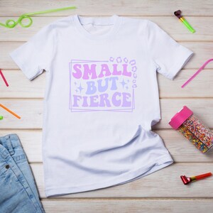 May include: White t-shirt with a pink and purple graphic that says "Small but Fierce" on a white wooden background.