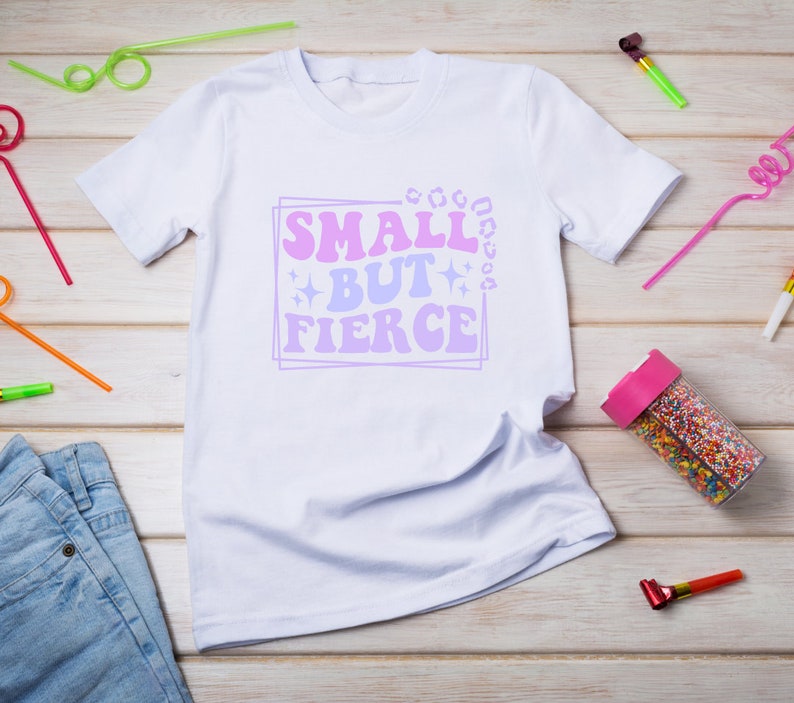 May include: White t-shirt with a pink and purple graphic that says "Small but Fierce" on a white wooden background.