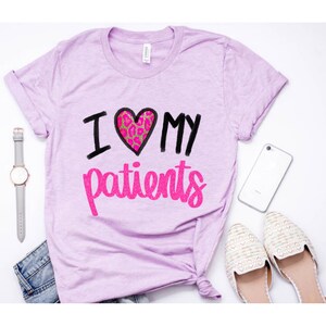 Puede incluir: Una camiseta de color morado claro con el texto "I love my patients" en letras negras y brillantes de color rosa. El coraz&oacute;n es rosa con un dise&ntilde;o de estampado de leopardo.