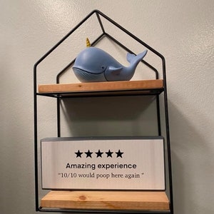 Puede incluir: Una estanter&iacute;a de metal negro con dos estantes de madera. Una figurita de ballena azul con un cuerno dorado se encuentra en el estante superior. Un letrero blanco con texto negro dice "Amazing experience" y "10/10 would poop here again".