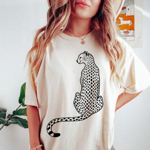 May include: Cream-colored t-shirt featuring a black and white cheetah illustration. The cheetah is depicted in a seated position with its tail curled. The shirt has short sleeves and a relaxed fit.