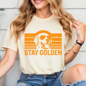 May include: Cream-colored t-shirt featuring a golden retriever wearing sunglasses, set against an orange sunset design. The phrase "STAY GOLDEN" is printed in bold, orange letters below the dog illustration. The shirt is made of a soft material.