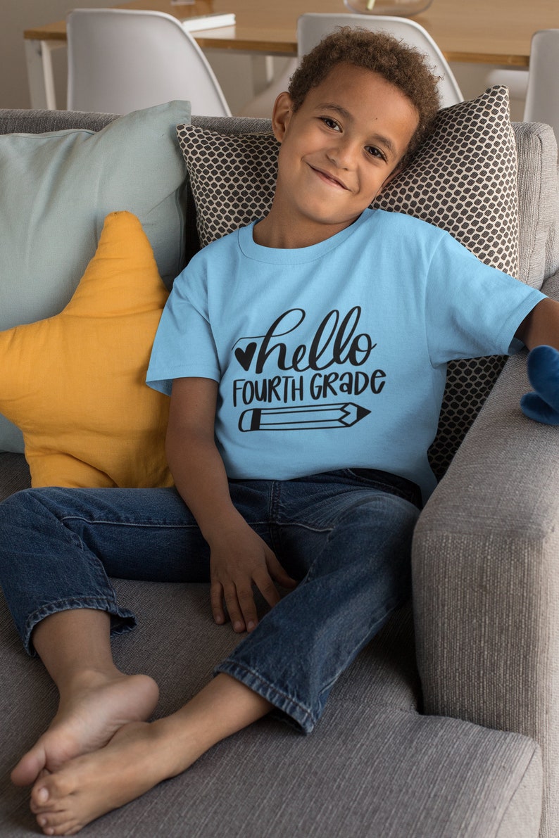 以下が含まれることがあります： 青いTシャツを着た子供が、グレーのソファに座っています。Tシャツには「hello fourth grade」という文字と鉛筆のイラストが描かれています。ソファには黄色の星型のクッションが置かれています。