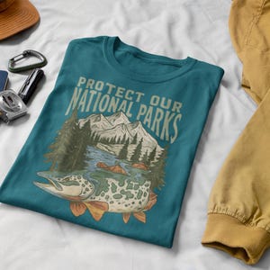May include: Teal t-shirt with the text "PROTECT OUR NATIONAL PARKS" and a graphic of a fish, mountains, and trees. Other items include a brown cap, a blue notebook, a silver carabiner, and a measuring tape.