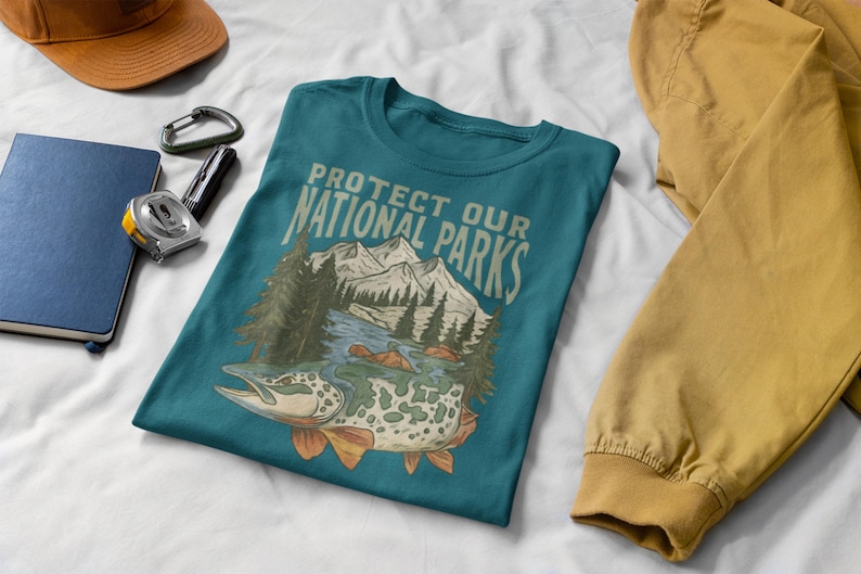 May include: Teal t-shirt with the text "PROTECT OUR NATIONAL PARKS" and a graphic of a fish, mountains, and trees. Other items include a brown cap, a blue notebook, a silver carabiner, and a measuring tape.
