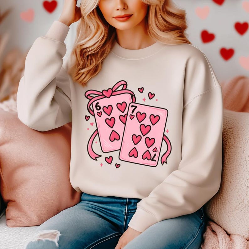 May include: A cream-colored sweatshirt featuring a graphic of two pink playing cards with heart symbols. The cards are tied with a pink ribbon and surrounded by small hearts. The cards show the numbers six and seven.