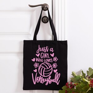 May include: Black canvas tote bag with pink text that reads "Just a girl who loves volleyball" with a pink volleyball graphic and hearts and flowers.
