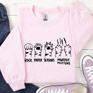 May include: Pink sweatshirt with a black graphic of cat paws and the text "Rock Paper Scissors Murder Mittens".